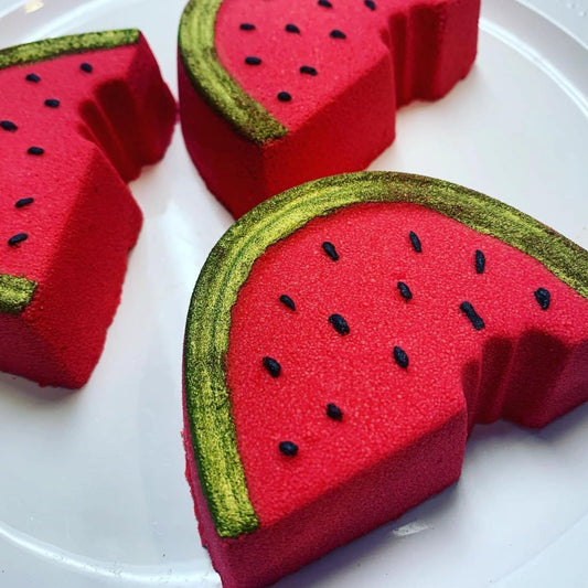Three red watermelon-shaped bath bombs with green rinds on a white plate.